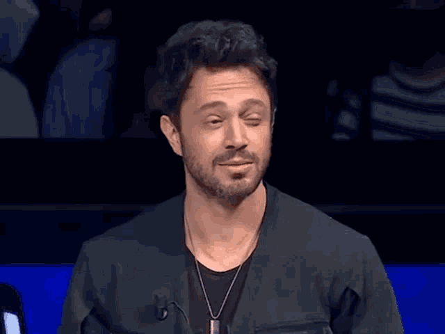 a man with a beard is wearing a black jacket and a necklace with the letter s on it