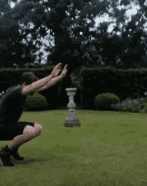 a man squatting on the grass with his arms outstretched in front of a statue