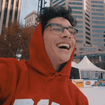 a man wearing glasses and a red hoodie laughs in front of a 40 hour sign