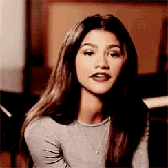 a woman with long hair is wearing a gray shirt and a necklace and looking at the camera .