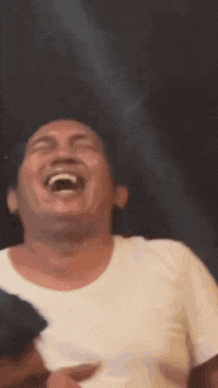 a man in a white shirt is laughing with his mouth open and his eyes closed .