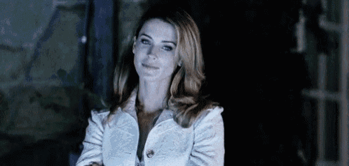 a woman in a white jacket is standing with her arms crossed in a dark room .