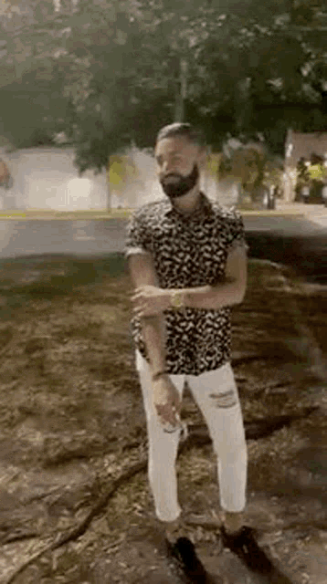 a man with a beard wearing a black and white shirt and white pants is standing on a sidewalk .