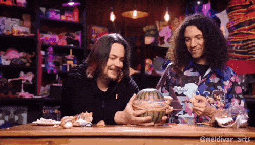 two men are sitting at a table and one is holding a watermelon with spaghetti on it
