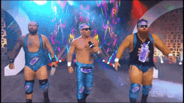 three men in blue wrestling outfits are walking on a stage with a microphone .