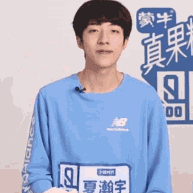 a young man wearing a blue new balance t-shirt is standing in front of a wall with chinese writing on it .