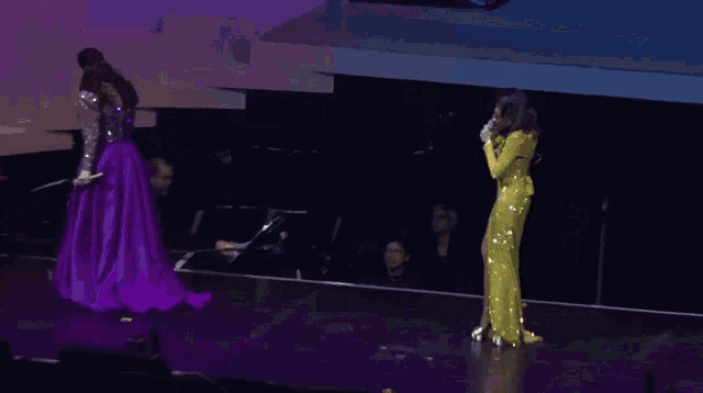 a woman in a purple dress stands next to a woman in a yellow dress on a stage