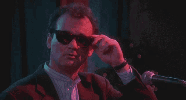 a man in a suit and sunglasses adjusts his sunglasses in front of a microphone