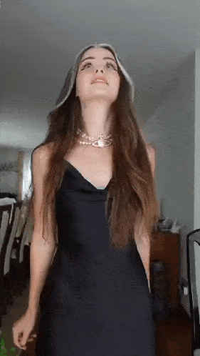 a woman in a black dress and pearl necklace is standing in a living room looking up .