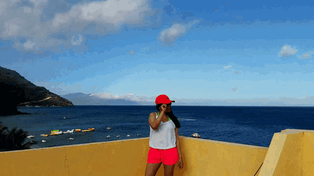a woman in a red hat and pink shorts stands on a balcony overlooking the ocean