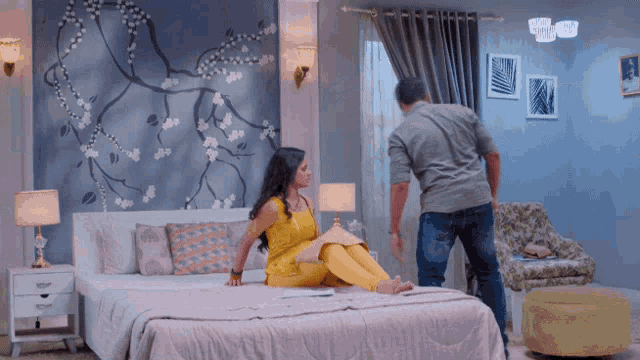 a woman in a yellow dress sits on a bed with a man standing next to her