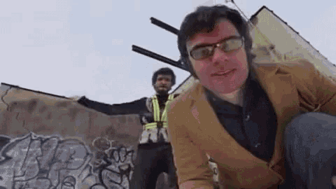 two men are standing next to each other in front of a wall with graffiti on it . one of the men is wearing glasses .