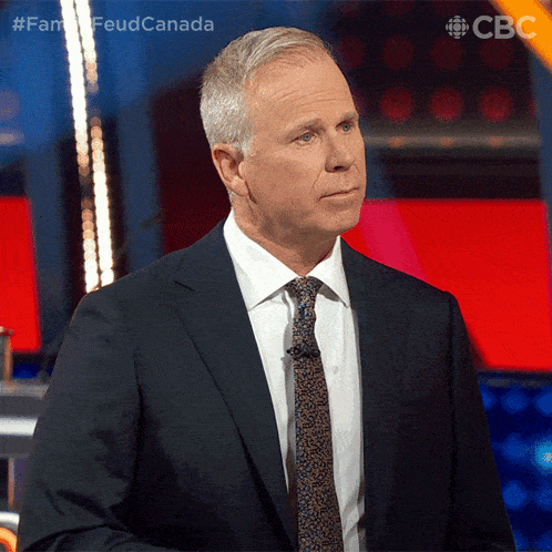 a man in a suit and tie stands in front of a microphone with the hashtag #famfeudcanada