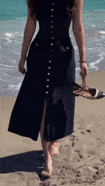 a woman in a black dress is standing on the beach holding her shoes