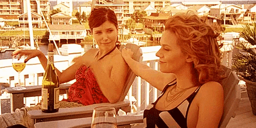 two women sitting on a balcony with a bottle of wine