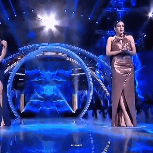 a woman in a long dress is standing on a stage with a circle in the background and the words zeudgifs on the bottom