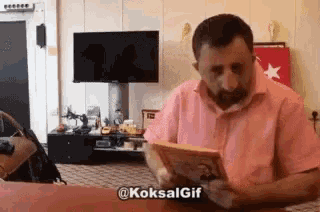 a man in a pink shirt is reading a book in front of a tv .