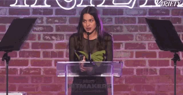 a woman stands at a podium with a microphone in front of a brick wall with the word variety on it