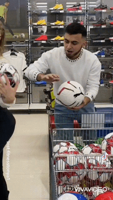 a man in a white sweater holds a soccer ball in front of a cart full of soccer balls