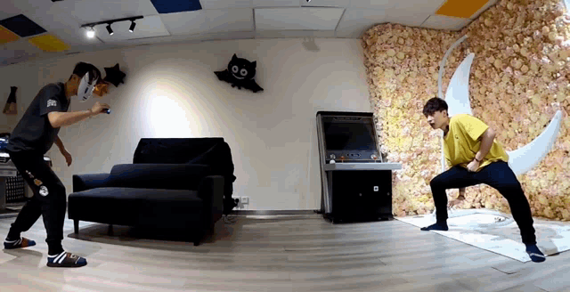 two men are playing a video game in a room with a couch and a wall of flowers