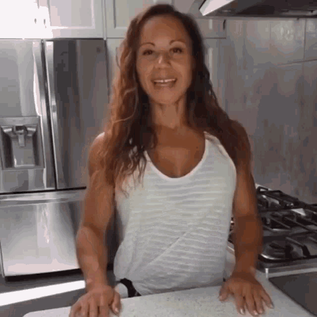 a woman in a white tank top is standing in a kitchen