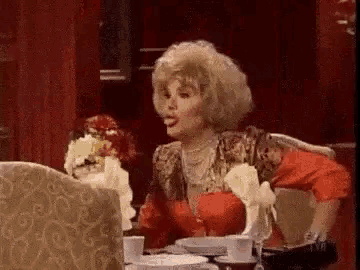 a woman is sitting at a table with plates and cups and talking to someone .