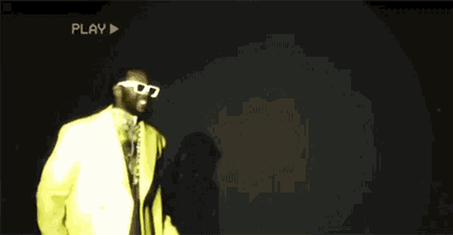 a man in a yellow suit and white sunglasses is dancing in front of a play button .