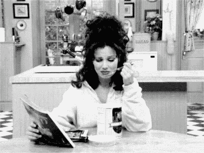 a black and white photo of a woman sitting at a table reading a magazine and eating food .