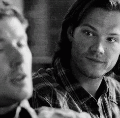 two men are sitting next to each other in a black and white photo . one of the men is pointing at the camera .