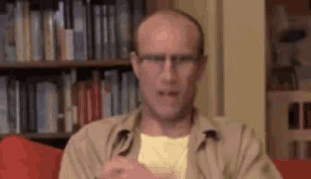 a bald man wearing glasses is sitting in front of a bookshelf .