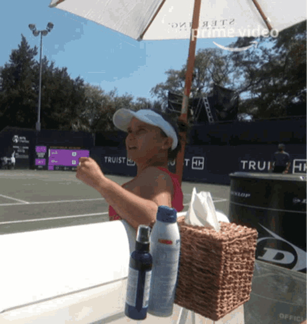 a woman sits under an umbrella in front of a tennis court sponsored by truist