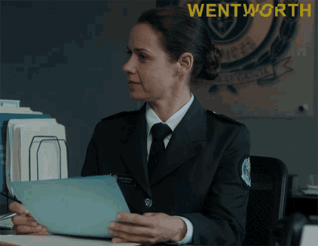 a woman in a police uniform is sitting at a desk holding a piece of paper in front of a wentworth sign