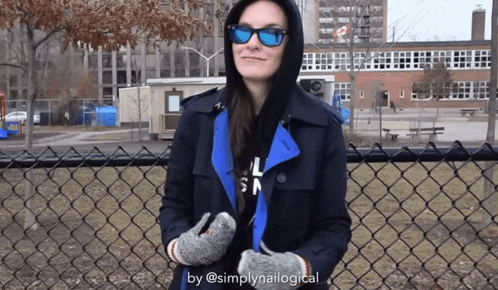 a woman wearing a hooded jacket and sunglasses is standing next to a chain link fence