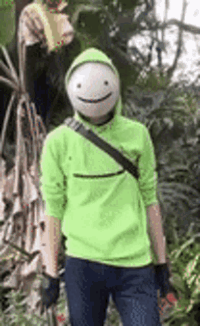 a man wearing a green hoodie and a white mask is standing in a forest .