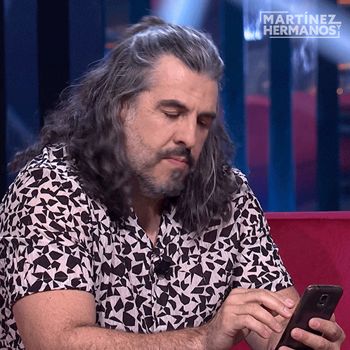 a man with long hair and a beard is looking at a cell phone with martinez hermanos written above him