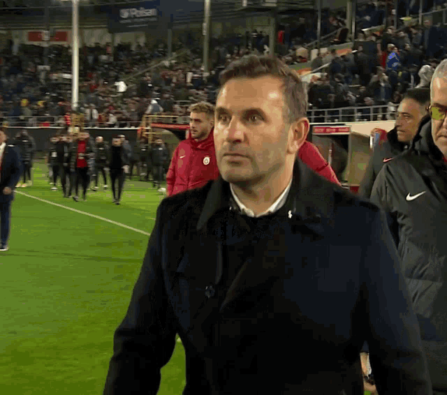 a man in a black jacket stands on a soccer field with a nike sign in the background