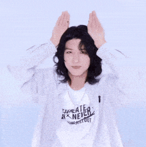 a young man with long hair is wearing a striped shirt and making a heart shape with his hands .
