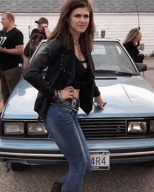 a woman stands in front of a blue car with a license plate that reads 4r4
