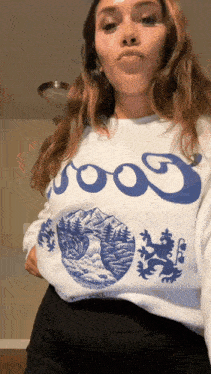 a woman is standing in a room wearing a white sweatshirt with a blue design on it .