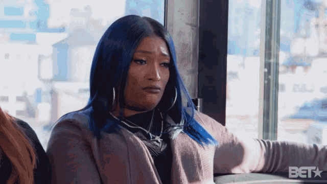 a woman with blue hair is sitting in front of a window with bet written on the bottom right