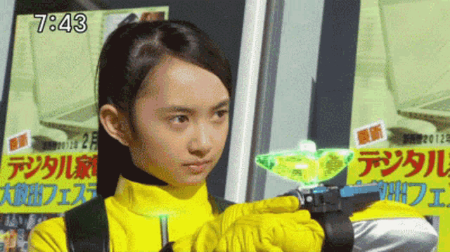 a girl in a yellow outfit is holding a green object in front of a sign that says " 7:43 "