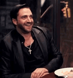 a man in a black jacket is sitting at a table with a plate of food .