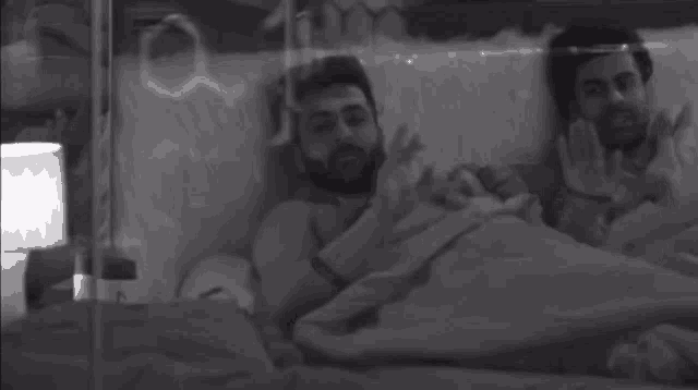 two men are laying in a bed under a blanket and talking to each other .