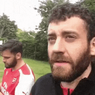 a man with a beard and a fly emirates shirt is standing next to another man .
