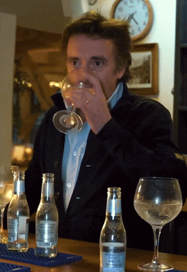 a man is drinking from a glass while sitting at a table with bottles of water and wine glasses