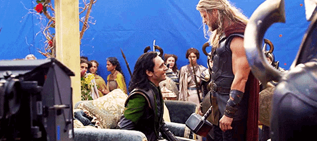 loki and thor are standing next to each other in a living room . thor is holding a hammer .