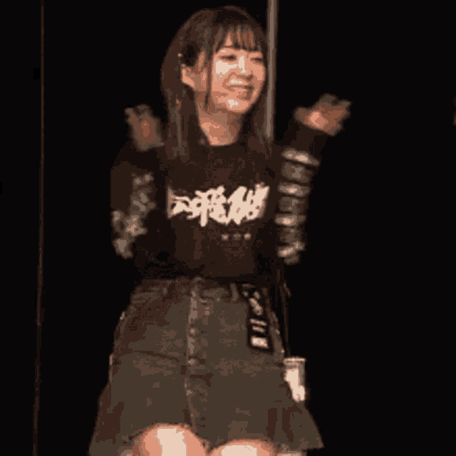 a woman in a black shirt and denim skirt is standing on a stage with her arms outstretched .