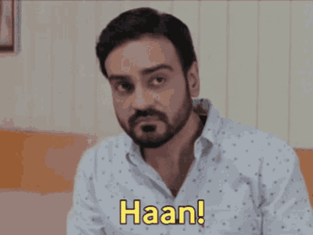 a man with a beard is sitting on a bed and making a funny face and saying haan !