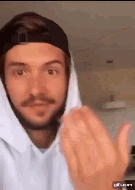 a man with a mustache wearing a hat and a white hoodie is giving a thumbs up .