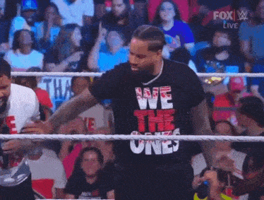 a man in a we the ones t-shirt is standing in a wrestling ring with a crowd behind him .